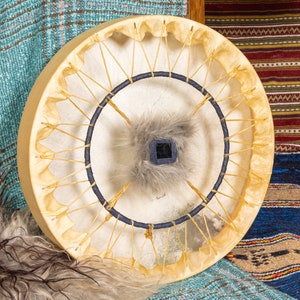 May include: A traditional Native American hand drum with a rawhide head, wooden frame, and a blue and white lacing pattern. The drum has a small, gray fur-covered center with a black square in the middle.
