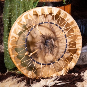 May include: A traditional Native American hand drum with a brown leather surface and a dark blue rope wrapped around the edge. The drum has a central hole with a brown fur patch in the center.