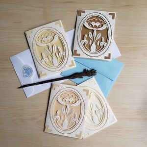 May include: Four white cards with a decorative floral design in an oval frame. The cards are on a light brown wooden surface. The floral design is cut out of the card and is white against a light yellow background.