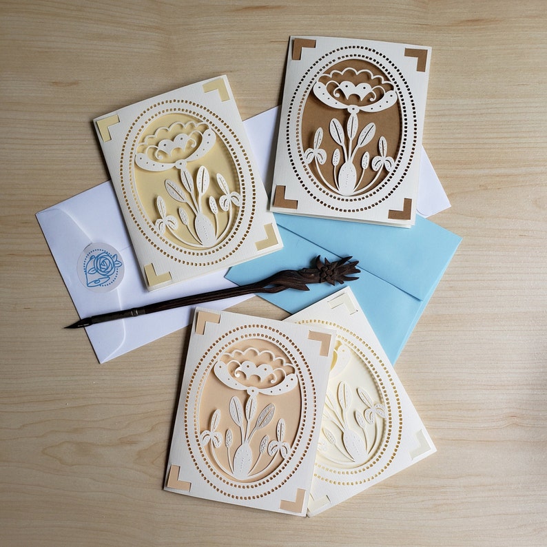 May include: Four white cards with a decorative floral design in an oval frame. The cards are on a light brown wooden surface. The floral design is cut out of the card and is white against a light yellow background.