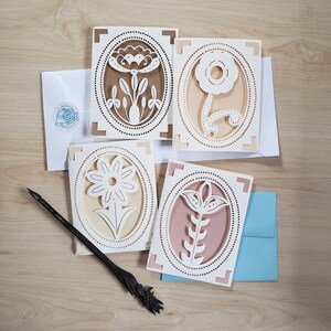 May include: Four white paper cards with intricate cut-out designs of flowers in oval frames. The cards are on a light brown wooden surface.