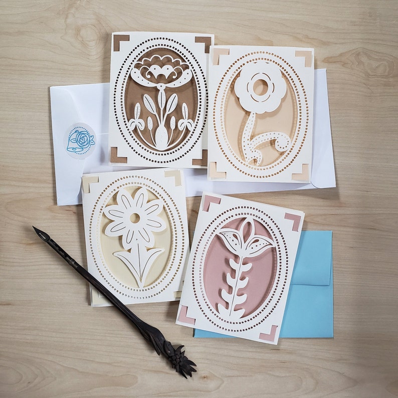 May include: Four white paper cards with intricate cut-out designs of flowers in oval frames. The cards are on a light brown wooden surface.