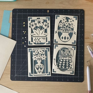 May include: Four intricately cut paper floral designs. Each card features a different flower arrangement in a vase or pot, with a teal background. The designs are set against a dark gray cutting mat, with craft tools and paper scraps visible.