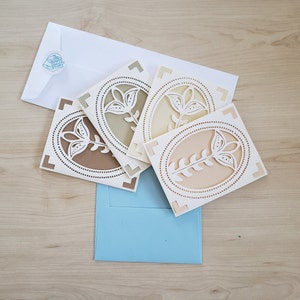 May include: Four white cardstock cards with a decorative floral design in a light brown, green, and pink color scheme. The cards are stacked on top of each other and are sitting on a blue surface.