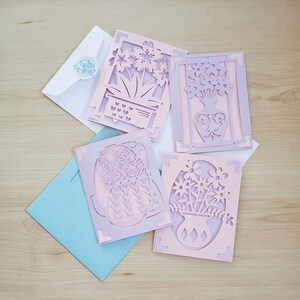 May include: Four laser-cut greeting cards with floral designs in shades of pink and lavender, each with a unique flower arrangement. The cards are accompanied by white and light blue envelopes, perfect for sending a thoughtful message.