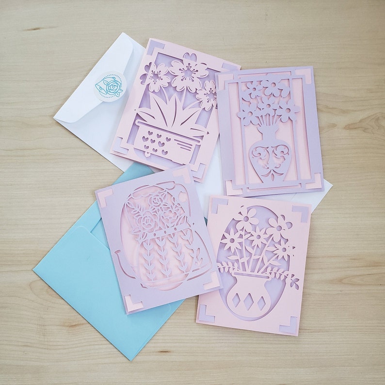 May include: Four laser-cut greeting cards with floral designs in shades of pink and lavender, each with a unique flower arrangement. The cards are accompanied by white and light blue envelopes, perfect for sending a thoughtful message.