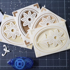 May include: Four white cardstock cards with a decorative, intricate, laser-cut design of a flower with a circle around it. The cards are on a black cutting mat with a white ruler and a blue tape dispenser.