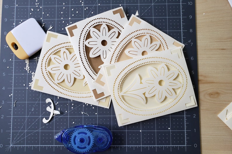 May include: Four white cardstock cards with a decorative, intricate, laser-cut design of a flower with a circle around it. The cards are on a black cutting mat with a white ruler and a blue tape dispenser.