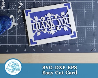 May include: A white card with a cut-out design of a floral wreath with the words "THANK YOU" inside. The card is on a blue background with a cutting mat.
