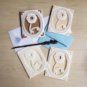 May include: Four handmade cards with a delicate floral design. Each card features a white paper flower with a brown or cream background. The cards are perfect for a special occasion.