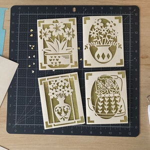 May include: Four laser-cut greeting cards with floral designs. Each card features a different flower arrangement, including flowers in a vase, a teapot, and a potted plant. The cards are made of light-colored cardstock with a gold glitter backing.