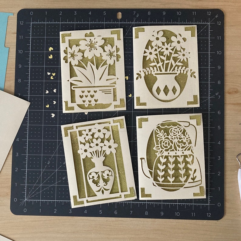 May include: Four laser-cut greeting cards with floral designs. Each card features a different flower arrangement, including flowers in a vase, a teapot, and a potted plant. The cards are made of light-colored cardstock with a gold glitter backing.