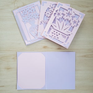 May include: A collection of handmade greeting cards with intricate floral cut-out designs. The cards feature a layered effect with light pink and lavender paper. One card is open, revealing a blank interior for a personalized message.