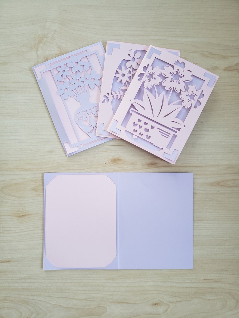 May include: A collection of handmade greeting cards with intricate floral cut-out designs. The cards feature a layered effect with light pink and lavender paper. One card is open, revealing a blank interior for a personalized message.