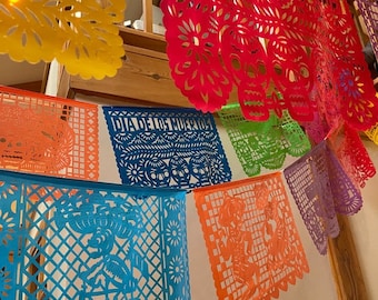 Puede incluir: Una colorida cadena de banderines de papel con diseños recortados, incluyendo calaveras y flores. Los banderines dicen "Dia de los Muertos".