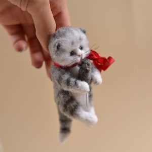 Peut inclure: Une petite figurine de chaton en feutre faite &agrave; la main avec des motifs de fourrure tigr&eacute;e grise et blanche. Le chaton a des yeux sombres, un nez rose et de longues moustaches, et porte un fin ruban rouge vif nou&eacute; autour du cou.