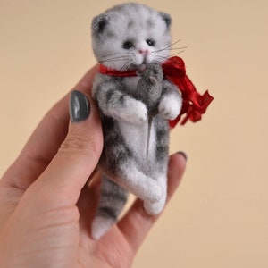 Peut inclure: Une petite figurine de chaton tigr&eacute; gris et blanc en laine feutr&eacute;e, dot&eacute;e d'yeux sombres et d'un nez rose. Le chaton porte un ruban rouge nou&eacute; autour du cou et tient une minuscule souris en feutrine gris fonc&eacute; entre ses pattes avant.