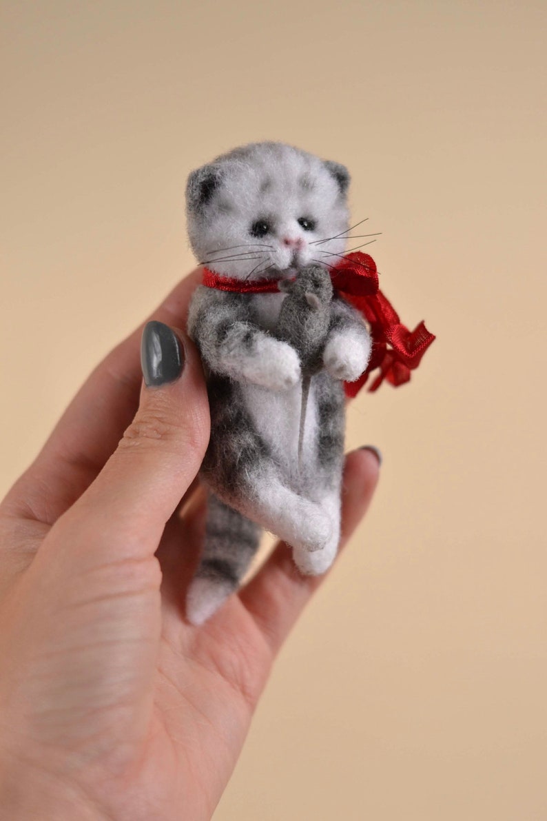 Peut inclure: Une petite figurine de chaton tigr&eacute; gris et blanc en laine feutr&eacute;e, dot&eacute;e d'yeux sombres et d'un nez rose. Le chaton porte un ruban rouge nou&eacute; autour du cou et tient une minuscule souris en feutrine gris fonc&eacute; entre ses pattes avant.