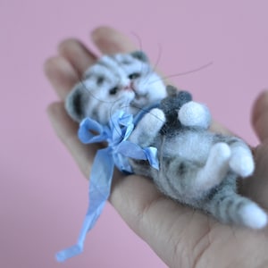 Peut inclure: Une petite figurine de chaton tigr&eacute; gris et blanc en laine feutr&eacute;e avec des rayures sombres, des pattes blanches et de longues moustaches. Le chaton est allong&eacute; sur le dos et porte un ruban bleu vif nou&eacute; autour du cou, avec une texture duveteuse.