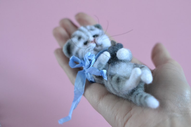 Peut inclure: Une petite figurine de chaton tigr&eacute; gris et blanc en laine feutr&eacute;e avec des rayures sombres, des pattes blanches et de longues moustaches. Le chaton est allong&eacute; sur le dos et porte un ruban bleu vif nou&eacute; autour du cou, avec une texture duveteuse.