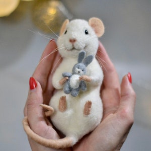 May include: A needle-felted white mouse figurine with pink inner ears and a long tail, holding a small grey felted rabbit toy. The mouse has black eyes, a pink nose, and long white whiskers, crafted with a soft, textured wool appearance.