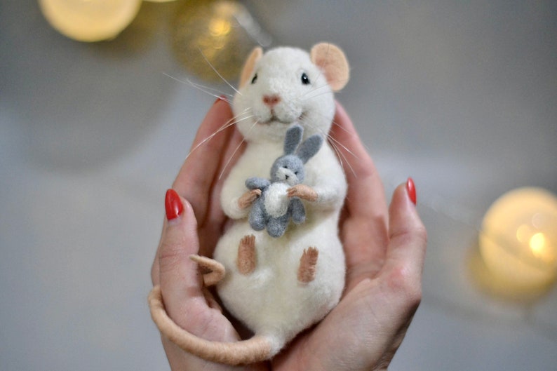 May include: A needle-felted white mouse figurine with pink inner ears and a long tail, holding a small grey felted rabbit toy. The mouse has black eyes, a pink nose, and long white whiskers, crafted with a soft, textured wool appearance.