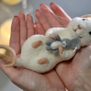 May include: A needle-felted white mouse figurine with pink ears and a long tail, holding a small grey and white felted rabbit toy. The mouse has black bead eyes, a pink nose, and long white whiskers, crafted from soft wool fibers.