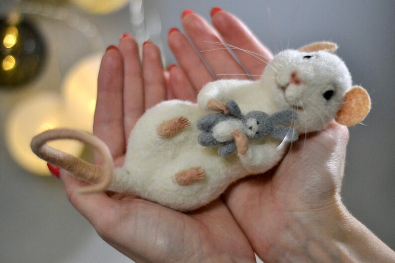 May include: A needle-felted white mouse figurine with pink ears and a long tail, holding a small grey and white felted rabbit toy. The mouse has black bead eyes, a pink nose, and long white whiskers, crafted from soft wool fibers.