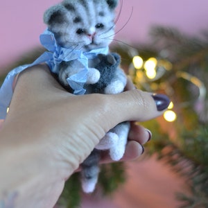 Peut inclure: Une figurine de chaton tabby gris et blanc en laine feutr&eacute;e avec des rayures fonc&eacute;es, des yeux noirs et de longues moustaches. Le chaton porte un ruban bleu clair nou&eacute; autour du cou et tient un petit objet gris fonc&eacute; entre ses pattes avant.