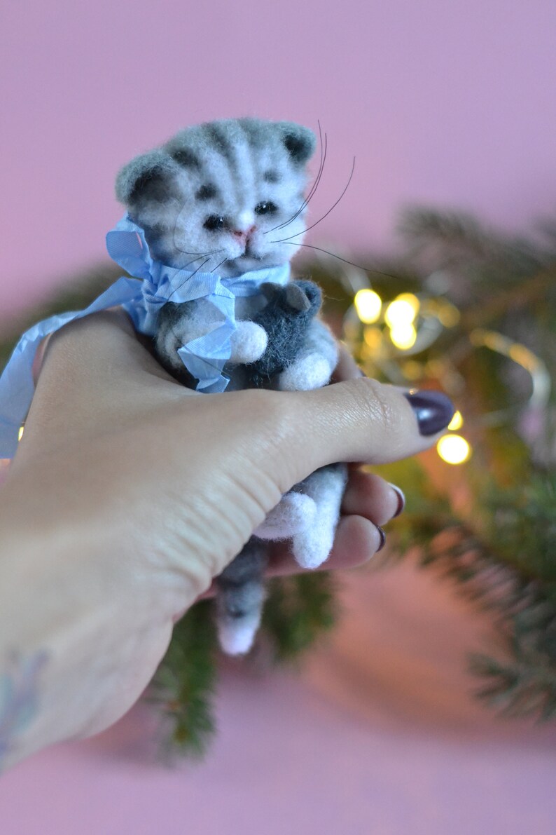 Peut inclure: Une figurine de chaton tabby gris et blanc en laine feutr&eacute;e avec des rayures fonc&eacute;es, des yeux noirs et de longues moustaches. Le chaton porte un ruban bleu clair nou&eacute; autour du cou et tient un petit objet gris fonc&eacute; entre ses pattes avant.