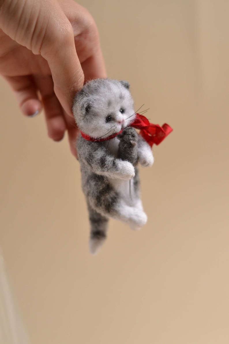 Peut inclure: Une petite figurine de chaton en feutre faite &agrave; la main avec des motifs de fourrure tigr&eacute;e grise et blanche. Le chaton a des yeux sombres, un nez rose et de longues moustaches, et porte un fin ruban rouge vif nou&eacute; autour du cou.