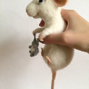 May include: A needle-felted white mouse figurine with pink inner ears and a long tail, holding a small grey felted mouse toy in its front paws. The mouse has dark eyes, a stitched nose, and thin whiskers, crafted from soft wool fibers.