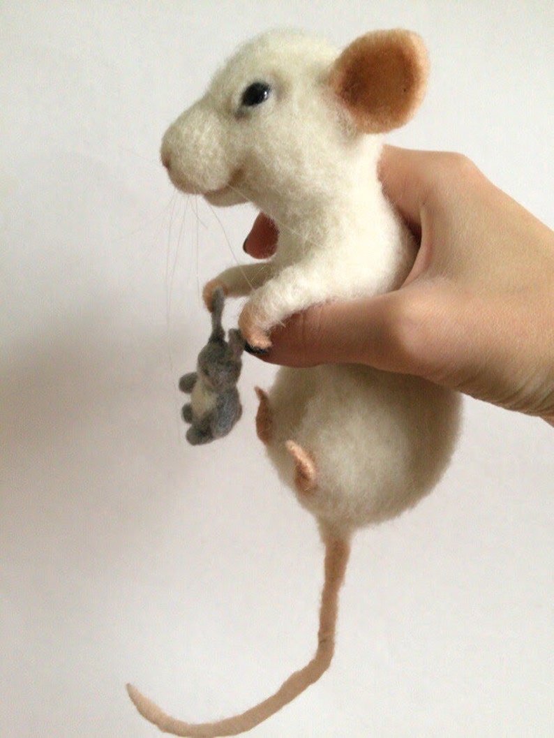 May include: A needle-felted white mouse figurine with pink inner ears and a long tail, holding a small grey felted mouse toy in its front paws. The mouse has dark eyes, a stitched nose, and thin whiskers, crafted from soft wool fibers.