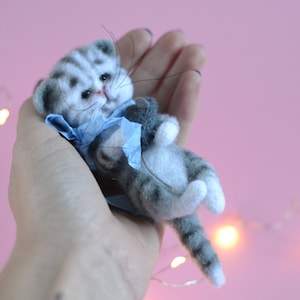 Peut inclure: Une petite figurine de chaton tigr&eacute; gris et blanc en laine feutr&eacute;e, avec des rayures fonc&eacute;es, des yeux noirs et de longues moustaches. Le chaton porte un accessoire ou un col en tissu bleu clair autour du cou et est en position allong&eacute;e.