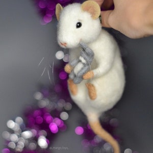 May include: A needle-felted white rat figurine with tan ears and feet, featuring black eyes and long whiskers. The rat is holding a small grey felted rabbit toy in its front paws. The text _margo_toys_ is visible near the bottom of the figure.