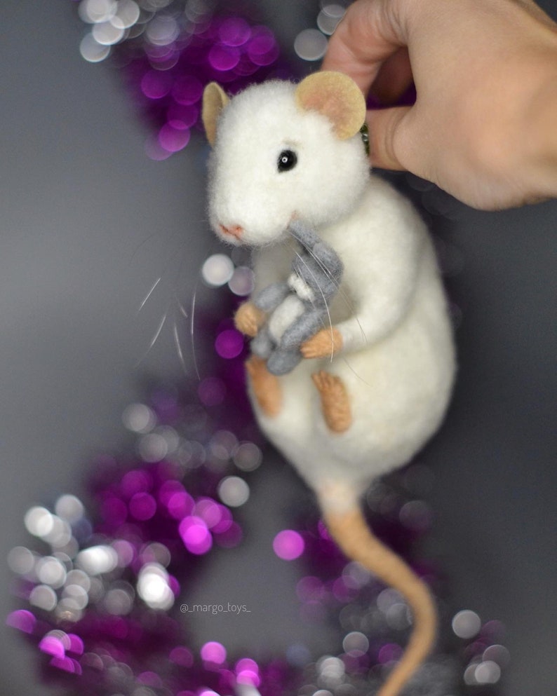 May include: A needle-felted white rat figurine with tan ears and feet, featuring black eyes and long whiskers. The rat is holding a small grey felted rabbit toy in its front paws. The text _margo_toys_ is visible near the bottom of the figure.