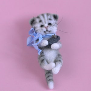 Peut inclure: Une figurine de chaton tigr&eacute; gris et blanc en laine feutr&eacute;e avec des rayures sombres, des yeux noirs et de longues moustaches. Le chaton porte un ruban bleu clair nou&eacute; autour du cou et tient un petit jouet poisson gris fonc&eacute; entre ses pattes.