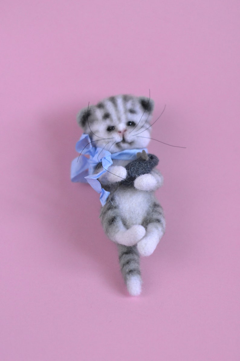 Peut inclure: Une figurine de chaton tigr&eacute; gris et blanc en laine feutr&eacute;e avec des rayures sombres, des yeux noirs et de longues moustaches. Le chaton porte un ruban bleu clair nou&eacute; autour du cou et tient un petit jouet poisson gris fonc&eacute; entre ses pattes.