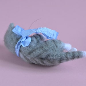 Peut inclure: Une figurine de chat tigr&eacute; gris en laine feutr&eacute;e avec des pattes blanches, allong&eacute;e sur le c&ocirc;t&eacute;. Le chat a un aspect textur&eacute; et duveteux et est orn&eacute; d'un ruban bleu clair nou&eacute; autour de son milieu, avec un petit n&oelig;ud sur le dessus.