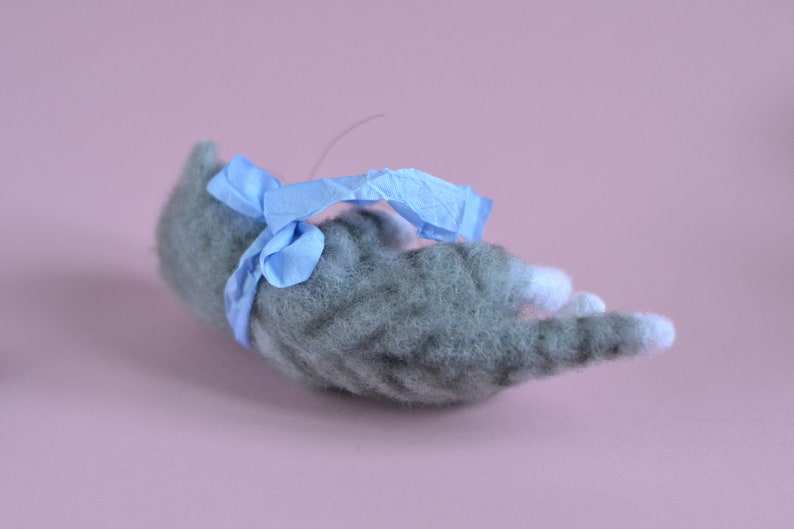 Peut inclure: Une figurine de chat tigr&eacute; gris en laine feutr&eacute;e avec des pattes blanches, allong&eacute;e sur le c&ocirc;t&eacute;. Le chat a un aspect textur&eacute; et duveteux et est orn&eacute; d'un ruban bleu clair nou&eacute; autour de son milieu, avec un petit n&oelig;ud sur le dessus.