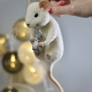 May include: A needle-felted white mouse figurine with pinkish ears and a long tail, holding a small grey stuffed animal in its front paws. The mouse has black bead eyes and thin, light-colored whiskers extending from its snout.