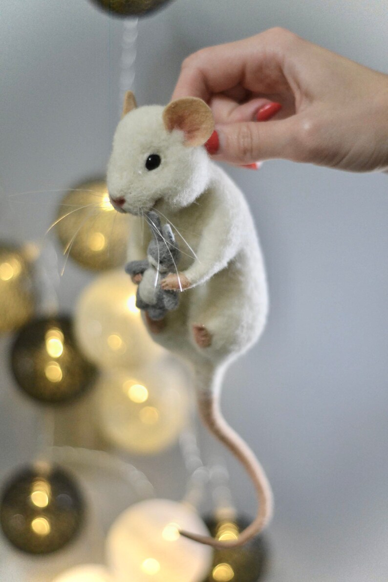 May include: A needle-felted white mouse figurine with pinkish ears and a long tail, holding a small grey stuffed animal in its front paws. The mouse has black bead eyes and thin, light-colored whiskers extending from its snout.