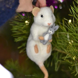 May include: A white needle-felted mouse figurine with tan ears, feet, and a long tail. The mouse has black eyes, a pink nose, and long whiskers, and it is holding a small, grey, felted rabbit toy. The text _margo_toys_ is visible at the bottom.