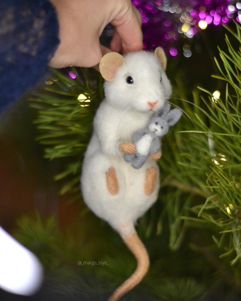 May include: A white needle-felted mouse figurine with tan ears, feet, and a long tail. The mouse has black eyes, a pink nose, and long whiskers, and it is holding a small, grey, felted rabbit toy. The text _margo_toys_ is visible at the bottom.