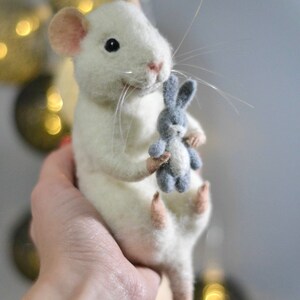 May include: A needle-felted white mouse figurine with pink-tinted ears and feet, featuring long white whiskers and black bead eyes. The mouse is holding a small, grey needle-felted rabbit toy in its front paws, showcasing a textured wool finish.