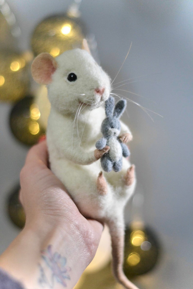 May include: A needle-felted white mouse figurine with pink-tinted ears and feet, featuring long white whiskers and black bead eyes. The mouse is holding a small, grey needle-felted rabbit toy in its front paws, showcasing a textured wool finish.