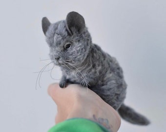 May include: A small, realistic needle-felted chinchilla figurine crafted from textured grey wool. The sculpture features large rounded ears, dark bead eyes, and long, thin white whiskers extending from its snout, capturing the likeness of a chinchilla.