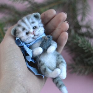 Peut inclure: Une petite figurine de chaton en laine feutr&eacute;e avec des rayures tigr&eacute;es grises et blanches, des yeux noirs et de longues moustaches. Le chaton porte un ruban bleu clair autour du cou et tient une petite souris en feutre gris dans ses pattes avant.