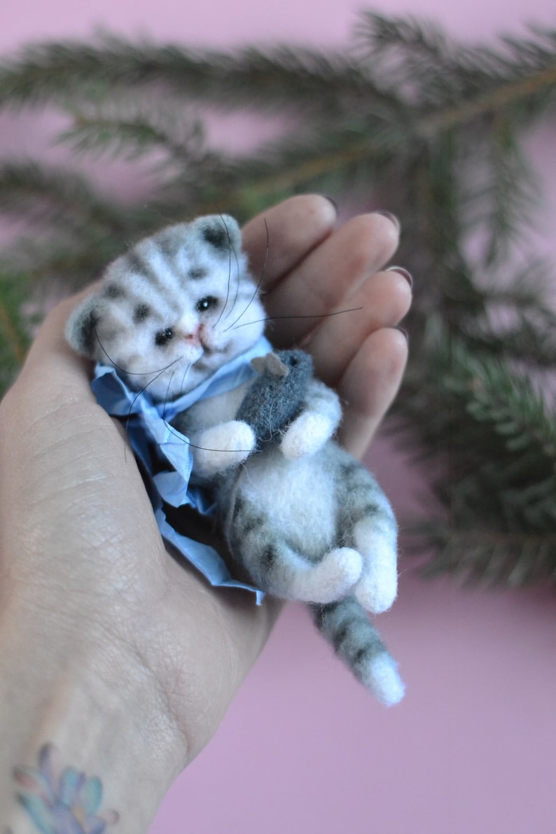 Peut inclure: Une petite figurine de chaton en laine feutr&eacute;e avec des rayures tigr&eacute;es grises et blanches, des yeux noirs et de longues moustaches. Le chaton porte un ruban bleu clair autour du cou et tient une petite souris en feutre gris dans ses pattes avant.