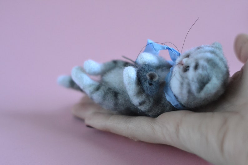 Peut inclure: Une petite figurine de chaton tigr&eacute; gris et blanc en laine feutr&eacute;e, allong&eacute;e sur le dos. Le chaton pr&eacute;sente des rayures gris fonc&eacute;, des pattes blanches et un ruban bleu clair nou&eacute; autour du cou. Il tient une minuscule souris en feutre gris fonc&eacute;.